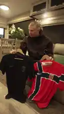 In a cozy indoor setting, Erling Haaland holds up two different Norway national team jerseys on hangers for comparison. He presents a black Nike jersey featuring the Norwegian Football Association crest alongside the team's iconic red home jersey, which displays the Nordic cross design. Dressed casually in a brown hoodie and blue sneakers, Haaland examines the garments closely, turning them slightly to show details like the Nike swoosh and sponsor logos. The scene captures a moment of personal style and national pride, showcasing the official football apparel. by @erling.haaland with Original Sound by erling.haaland