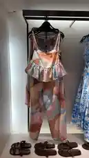 A curated display of a modern women's two-piece outfit featuring a ruffled crop top and wide-leg pants in a soft, abstract color-block pattern. The ensemble is styled with brown leather sandals, presented on a sleek black hanger against a minimalist retail backdrop. This fashion-forward look reflects contemporary trends from Rawaiah Al Sharqiyah, a prominent brand based in Taif, Saudi Arabia, known for its elegant and culturally inspired designs. by @ru.ab71 with original sound
