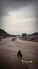 This scene captures a dramatic moment where a desert road has been transformed into a rushing river due to flash flooding. Numerous white SUVs and trucks are seen carefully driving through the deep, muddy water, while a group of people, including women in traditional black abayas, stand on the dry bank observing the situation. A large, rocky hill dominates the background under a heavy, overcast sky, emphasizing the severity of the weather event. by @karim.alsaleh with يجي مطره🌧️ by Contains: صوت مطر ياجي - عائلة موكا ممادو