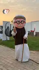 A cheerful elderly mascot character, representing فريق رسالة سلام التطوعي, poses outdoors during a volunteer event at golden hour. Dressed in traditional attire with a walking stick, the mascot makes a peace sign, embodying the group's message of goodwill. The scene captures community spirit and positive outreach in a warm, inviting atmosphere. by @peace.message with original sound