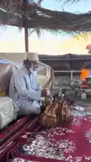 An elderly man in traditional Saudi attire prepares and serves Arabic coffee over an open fire under a palm-thatched roof. This authentic cultural moment captures the ritual of making qahwa, often accompanied by dates, set against a scenic desert backdrop. by @azozi.5562 with Original Sound by azozi.5562