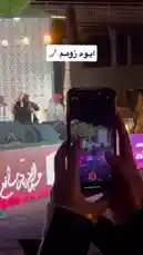 This scene-by-scene summary details a real-time recording of a live event. A person holds up their smartphone, using the camera app to film a stage performance. On the screen, performers in traditional attire are visible, along with a large decorative backdrop and stage lighting. The focus shifts subtly, highlighting the act of documenting a cultural gathering, likely a 'Jalsah' event, emphasizing the importance of capturing memories through mobile technology. by @jalsahevent with jalsahevent by Original Sound