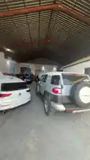 This walkthrough video provides a clear view of the interior of a bustling auto repair shop. Several cars, including a white Volkswagen Golf and a silver Toyota FJ Cruiser, are parked inside the spacious garage with its distinctive corrugated metal roof. Some vehicles have their hoods open, suggesting they are undergoing maintenance or repairs. The scene captures the typical environment of a professional mechanic's workspace. by @hawkar_service with Original Sound by hawkar_service