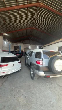 This walkthrough video provides a clear view of the interior of a bustling auto repair shop. Several cars, including a white Volkswagen Golf and a silver Toyota FJ Cruiser, are parked inside the spacious garage with its distinctive corrugated metal roof. Some vehicles have their hoods open, suggesting they are undergoing maintenance or repairs. The scene captures the typical environment of a professional mechanic's workspace. by @hawkar_service with Original Sound by hawkar_service
