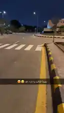 A first-person view of a quiet city street at night. The camera moves forward along a road marked with a yellow line and a black-and-yellow curb. A pedestrian crosswalk is visible ahead, and a dark car drives past in the distance under the glow of streetlights. by @khllo17 with Original Sound by khllo17