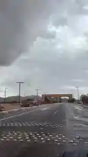 A first-person perspective journey down a wet road leading toward a large stone archway in a desert landscape. Dark, heavy clouds dominate the sky, suggesting an impending storm. The road features reflective markers and is flanked by streetlights and sparse vegetation. This atmospheric scene captures the mood of a dramatic weather event in a remote, arid region. by @azozi.5562 with ديار ⛈️🤍 . by Azozi.5562