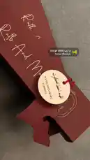 This close-up shot reveals the elegant packaging of 'Ratb And More,' a brand known for its sophisticated aesthetic. The deep red box features stylish white script lettering and a circular tag with both English and Arabic text, including the brand name and the date '22/06/2023.' A decorative cutout adds a modern touch to the design. The scene is set against a neutral grey background, emphasizing the product's premium feel. by @om.louraa with يا رب احفظ كويتنا by Bader Alnufais