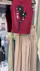 A close-up showcase of a fashionable women's outfit set. The ensemble features a vibrant red sleeveless tank top adorned with prominent black floral appliqués and a delicate drawstring detail. Paired with it are elegant, light beige wide-leg pants with a matching drawstring waistband. The clothing is displayed on a hanger within a well-lit retail environment, suggesting a new arrival or a featured look from the 'كود اناقتي' collection. by @code.a3 with original sound