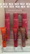 A static shot showcases the new Blushing Boutique lip gloss collection. Three vibrant shades—orange, red, and brown—are displayed in clear tubes with unique charms, set against their pink packaging. The background features a large graphic of lips and the text 'NEW NEW NEW,' creating a fresh and exciting reveal for the brand's latest beauty products. by @bx301a with 🔫🔫🔫🔫🔫🔫🔫🔫🔫 by Contains: Charger - Triangle des bermudes