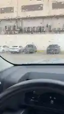 A first-person perspective from inside a car shows a rainy day commute. The driver navigates through wet streets, with raindrops heavily covering the windshield and side windows. The view includes passing parked cars and a building with multiple air conditioning units. A male voice speaks in Arabic, expressing relief at arriving at his destination despite the downpour, creating a relatable moment for anyone who has driven in bad weather. by @guruofmarketing with Original Sound by guruofmarketing