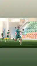 A dynamic soccer training moment captured in real-time shows a player from نادي الروضة السعودي executing a powerful kick while airborne. The athlete, wearing a green jersey with yellow accents, leaps into the air to strike the ball mid-flight during a drill on a well-maintained pitch. Other teammates in matching kits are visible in the background, suggesting an organized practice session. The stadium's tiered seating, partially filled with orange and green seats, adds context to the professional training environment. by @alrawdahclub with original sound
