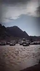 A person wades through a fast-flowing river in a rugged mountain landscape, surrounded by parked off-road vehicles. The dramatic scenery, with steep rocky peaks under a cloudy sky, sets the stage for an adventurous outdoor expedition. by @karim.alsaleh with ياعمليات ياعمليات 💔😂 . ‏ by sn: z-ws5