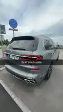 This video offers a stylish rear view of a silver BMW M760Li xDrive parked on a city street under a cloudy sky. The camera slowly pans around the vehicle, highlighting its elegant design, dual exhausts, and distinctive taillights. A 'Sjukt smutt slåde' watermark suggests a personal or social media context. The surrounding environment includes a 'CHOKOLAD' sign and urban infrastructure, placing the scene in a modern European setting. by @tershine with original sound