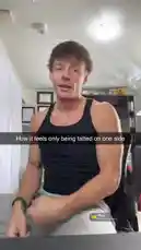 In this scene, Dylan Terryberry is seated in his room, reacting to a new music release. He is wearing a black tank top and gray sweatpants. The room has a bookshelf in the background, along with various framed pictures and a plaid blanket on a couch. The lighting is bright, coming from a ceiling light. Dylan appears engaged and expressive as he watches the content. by @datloading with Mine by Tink (feat. G Herbo)