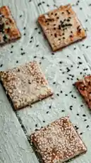 Watch as square sesame crackers are beautifully arranged on a rustic wooden surface. These golden-brown treats are generously sprinkled with white and black sesame seeds, creating a visually appealing texture. Perfect for snack lovers and food enthusiasts exploring simple, homemade recipes. by @moladone with original sound
