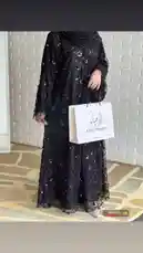 A stylish individual showcases a long black lace abaya adorned with floral patterns, paired with a matching hijab. They hold a white paper shopping bag branded 'Khalil Abegha' and wear sparkly heels, standing against a white brick wall. The scene is accompanied by Arabic pop music. by @mohammed_aldrop with انا وقلبي نتفق❤️ by ❤️❤️❤️❤️❤️❤️❤️