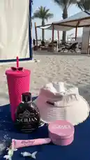 A stylish beach setup featuring a pink tumbler, a white bucket hat, a bottle of Sicilian tanning lotion, and a jar of Cosmo beauty cream. The serene sandy beach backdrop with palm trees and lounge chairs creates a perfect atmosphere for relaxation and self-care. by @bx301a with 🌴🌴🌴🌴🌴 by Contains: HOT UPTOWN - Camila Cabello, Drake