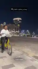 A young man on a yellow bicycle stops on a city sidewalk at night. A woman approaches him and they exchange a small gift, sharing a smile. The illuminated city skyline serves as a backdrop for this brief, friendly interaction. by @sequence-coffee with لقطة سعيـدة❤️ by .