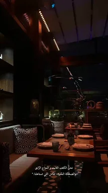 This video captures the serene atmosphere of a dimly lit restaurant or lounge area during the evening. The setting features wooden tables set for dining, modern chairs, and patterned cushions on a bench. Warm, ambient lighting from the ceiling and decorative lamps creates a cozy mood. Through a large glass wall, a dark outdoor scene with some greenery is visible. Throughout the clip, traditional Arabic music plays, enhancing the tranquil and intimate ambiance. by @batoolmakeup1 with الليل ما يحلا الا بجلاسُه🎻 by .