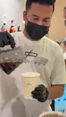 A close-up shot captures a barista from Sequence Coffee demonstrating their pour-over coffee technique. Wearing a branded white t-shirt, black gloves, and a face mask, the barista carefully pours hot water from a glass kettle into a paper cup. The focus is on the precise and skillful motion required to brew a high-quality cup of coffee. by @sequence-coffee with Original Sound by sequence-coffee
