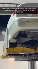 This nighttime footage provides a behind-the-scenes look at the professional car wash service 'Magsa Al Arba Qatir'. It begins with a close-up of a silver sedan being thoroughly sprayed under a lift, then pans out to reveal the full scale of the operation. Workers in red shirts can be seen actively cleaning vehicles under bright lights, while other cars wait their turn under a large yellow canopy. The modern facility features prominent Arabic signage, showcasing its branding and operational efficiency. by @wash.4drops with Original Sound by wash.4drops