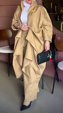 This video showcases a full-length fashion look from Artal Fashion أرتال. A model presents a stylish ensemble featuring a long, structured khaki trench coat with dramatic ruffled layers over matching wide-leg trousers. The outfit is paired with a white turtleneck, black pointed-toe heels, and a textured black clutch adorned with a colorful charm. The setting is a modern indoor space with wooden chairs, highlighting the contemporary design and elegant silhouette of the clothing. by @artal.street with original sound