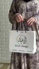 This stylish video features Mohammed from Khial Abaya presenting a beautiful, long-sleeved abaya dress with a subtle grey floral pattern. He models the elegant outfit, which includes a black hijab and sparkly shoes, while holding a branded shopping bag for the 'Khial Abaya' brand. The setting is a modern interior with white brick walls, highlighting the contemporary design of the garment. by @mohammed_aldrop with Original Sound by mohammed_aldrop