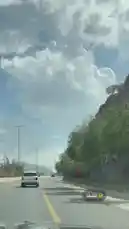 This captivating footage captures a journey along a modern highway winding through a mountainous region. The viewer is treated to stunning views of towering, rocky cliffs adorned with lush greenery and vibrant red flowering bushes. Above, a vast sky filled with massive, sculptural cumulus clouds creates a breathtaking backdrop, suggesting either early morning or late afternoon light. The road itself is wide and well-maintained, featuring clear yellow lane markings and tall, sleek streetlights lining one side. A white SUV is visible ahead, traveling in the same direction, providing a sense of scale and movement. The overall atmosphere is one of serene travel and natural beauty, showcasing a picturesque route perfect for a scenic drive. by @om.louraa with original sound