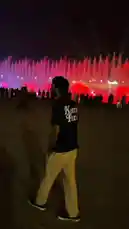 A man in a Kenzo Paris t-shirt watches a spectacular nighttime fountain light show. The water jets dance in sync with vibrant blue, red, yellow, and purple lights, creating a mesmerizing spectacle for a crowd of onlookers. by @moazzam with Original Sound by moazzam