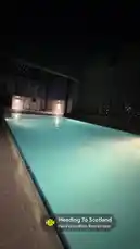 A serene night view of a well-lit rooftop swimming pool, capturing the tranquil ambiance of a modern urban retreat. The calm water reflects the soft glow of surrounding lights, creating a peaceful atmosphere. This moment is part of a travel vlog titled 'Heading To Scotland,' suggesting a journey or transition to a new destination. by @dsunmusic with Heading To Scotland by Harshavardhan Rameshwar