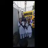 A group of young students from ITH School stands in formation outdoors, dressed in matching white uniforms with black sashes. They perform a synchronized hand gesture routine in front of a large yellow banner displaying the text 'WHERE LIGHT' and a cartoon character. An adult woman in a headscarf supervises them. The performance takes place on a paved area next to a wall topped with barbed wire, suggesting a school courtyard or event space. by @ithschool with Original Sound by ithschool