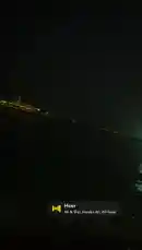 This nighttime video captures a serene waterfront view, panning across a dark body of water reflecting the lights of a distant city skyline and a brightly illuminated bridge. The gentle movement of the water creates shimmering reflections, while a music player overlay indicates the track 'Heer' by Ali & Shijr, Haider Ali, Ali Raza is playing. The overall atmosphere is calm and peaceful. by @moazzam with Heer by Ali & Shjr, Haider Ali, Ali Raza