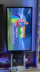 This video provides a view of Navneet's television setup, where a sports broadcast celebrating 'INDIA CHAMPIONS' is being displayed. The screen shows a player holding the Indian flag, followed by a spectacular fireworks display over a stadium at night. The surrounding entertainment center features decorative items like flowers and a black figurine, all illuminated by colorful LED strip lights. by @im.navnit with Original Sound by im.navnit