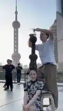 Witness a captivating street performance in Shanghai where a woman demonstrates incredible balance, keeping a large, tower-like structure perfectly balanced on her head. The scene unfolds on a bustling city terrace with the iconic Oriental Pearl Tower illuminated in the background, transitioning from day to night. This mesmerizing display of skill and artistry draws the attention of passersby. by @maddi_op with maddi_op by Original Sound