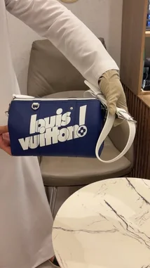 A close-up showcase of a blue Louis Vuitton pouch bag with white branding and strap. A person in a white outfit displays the bag from multiple angles, highlighting its design and size. An Arabic voiceover describes the product as 'nice' and states its price is 7,500. by @lussowatches with Original Sound by lussowatches
