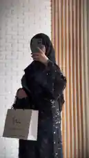 A woman models a stunning black abaya adorned with intricate silver floral embroidery and sequins. She accessorizes with a matching hijab, rings, and a white shopping bag from 'Khal Abya'. The stylish look is presented against a modern white brick wall background. by @mohammed_aldrop with Original Sound by mohammed_aldrop