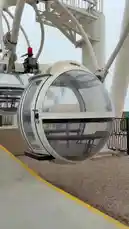 This video provides a first-person perspective from inside a large, transparent gondola of a Ferris wheel, numbered '16'. The cabin is shown being loaded at a station before ascending to reveal a sweeping view of a beach below. The scene captures a line of palm trees along the shore, waves crashing onto the sand, and several parked cars, all under an overcast sky. The camera pans across the scenery, offering a unique vantage point of the coastal landscape. by @nowaira-11 with وبطير من كتر الفرح by ❤️❤️💓💗💗💖💝💝💞💘💘