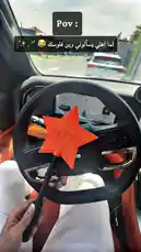 This first-person perspective video captures a unique interaction inside a modern luxury car. The driver holds a distinctive orange star-shaped wand with '4B' printed on it. Outside the vehicle, a smiling delivery person wearing gloves and a branded hoodie approaches the car window, holding a tablet and a small white package. The setting appears to be a sunny day at a commercial or entertainment complex, suggesting a contactless delivery service. The scene highlights the blend of technology, personalization, and premium experience. by @th4eb with Original Sound by th4eb