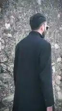 A stylish man, identified as Moazzam Ishaq, walks confidently past a weathered stone wall. He is wearing a long black coat over a light-colored collared shirt and distinctive aviator sunglasses. The camera follows him from behind before he turns to face the lens, showcasing his modern look and beard. by @moazzam with Original Sound by moazzam