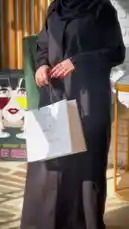 A stylish look showcasing a modern black abaya with feather-like embroidery, paired with a chic hijab and red heels. The model displays the outfit from multiple angles, highlighting its elegant design and flow. She accessorizes with a white shopping bag from 'Khil Abaya' and a small red handbag, presenting a fashionable and contemporary Islamic modest wear ensemble. by @mohammed_aldrop with Original Sound by mohammed_aldrop