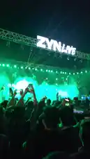 A behind-the-scenes look at the electrifying atmosphere of a ZYN LIVE concert. The crowd is fully engaged, recording the performance on their phones under vibrant green stage lights and atmospheric smoke. A fan in a light-colored t-shirt stands out, capturing the moment from the audience perspective. by @moohid_77 with Original Sound by moohid_77