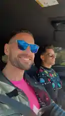 Duke Sole enjoys a sunny day drive with a friend, showcasing his vibrant style. He sports a bright pink shirt under a silver jacket and blue mirrored sunglasses. His passenger wears a colorful Pac-Man sweater and round shades. The pair smiles and laughs together, creating a fun and stylish atmosphere. by @duke_sole with original sound