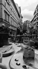 This black-and-white time-lapse captures the serene ambiance of a European city street café. A slice of cake sits on a plate in the foreground as the scene unfolds. A bird lands on the table, briefly interacting with the dessert before flying away. In the background, people sit at outdoor tables, enjoying their meals under a cloudy sky. The timeless architecture of the surrounding buildings frames this peaceful moment of urban life. by @two with ربنا فاغفر لنا ذنوبنا - قران كريم by طريق القران