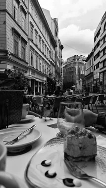 This black-and-white time-lapse captures the serene ambiance of a European city street café. A slice of cake sits on a plate in the foreground as the scene unfolds. A bird lands on the table, briefly interacting with the dessert before flying away. In the background, people sit at outdoor tables, enjoying their meals under a cloudy sky. The timeless architecture of the surrounding buildings frames this peaceful moment of urban life. by @two with ربنا فاغفر لنا ذنوبنا - قران كريم by طريق القران