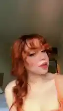 A young woman with vibrant orange hair styled in a braid and bangs is featured in a close-up shot. She wears a matching orange tank top and has full makeup on, including winged eyeliner and blush. She looks directly at the camera, speaking and making various expressions. The background appears to be a bedroom with a bed visible. An upbeat, rhythmic song with a male vocalist singing in a foreign language plays throughout the clip. by @merlinblok3 with Original Sound by merlinblok3