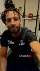 This static image captures fitness influencer Jack Donnovan in a gym setting. He is looking directly at the camera with a slight smile. He has dreadlocks styled in a bun and is wearing a black t-shirt with white and red graphics. A text overlay on the image reads 'On a fit 💯'. In the background, gym equipment, including a weight rack with red pads, is visible. by @jackdonnovan with Original Sound by Contains: I FEEL GOOD - Mike Teezy
