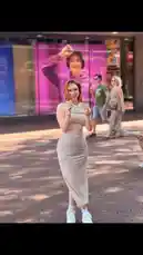 A young woman poses confidently on a brick-paved city street, showcasing a stylish striped dress paired with white sneakers. She holds a lollipop, adding a playful touch to her look. In the background, a large advertisement featuring a person in a purple hoodie is visible on a storefront window, while other pedestrians walk by under the bright sun. by @merlinblok3 with Yangın Yeri by Elif Buse Doğan