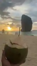 오리지널 사운드 포함 @hamza_sheikh8 님이 만든 A serene sunset unfolds at the iconic Maya Bay, with a hand holding a fresh coconut drink in the foreground. The golden sun dips below the horizon, casting warm light across the beach and sea. In the distance, the towering limestone karst known as Koh Lanta stands majestically against the sky. A few people can be seen enjoying the tranquil moment, capturing memories under the vast, colorful sky.