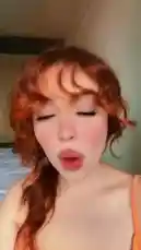 A close-up, selfie-style video featuring a young woman with vibrant red hair styled in loose waves and bangs. She wears a bright orange spaghetti strap top and has full makeup on, including winged eyeliner and pink blush. The setting appears to be a bedroom with a bed visible in the background. She moves her head and body rhythmically, lip-syncing to a Turkish pop song playing in the background. The overall aesthetic is bright, casual, and focused on personal style. by @merlinblok3 with Original Sound by merlinblok3
