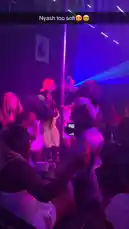 A woman in a grey bodysuit dances provocatively on a rotating pole in a crowded nightclub. The scene is illuminated by flashing blue, red, and green lights, creating an energetic party atmosphere. Other guests watch and dance around her. by @be-sassyish with Original Sound by be-sassyish